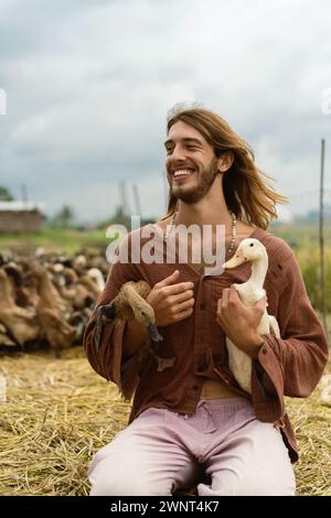 Un homme aux cheveux longs souriant tient des canards dans ses mains. Banque D'Images