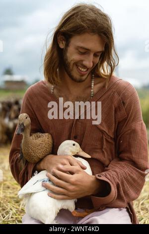 Un homme aux cheveux longs souriant tient des canards dans ses mains. Banque D'Images