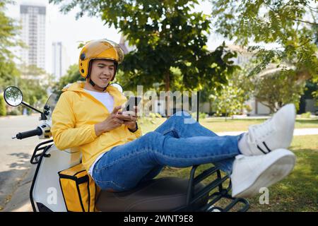 Courrier souriant assis sur un scooter, se reposant entre les manèges, envoyant des SMS à des amis Banque D'Images