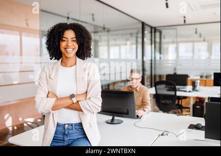 Une femme afro-américaine confiante avec un sourire radieux se tient les bras croisés. L’arrière-plan révèle un collègue concentré sur son écran, mettant en évidence un environnement de travail professionnel et dynamique Banque D'Images