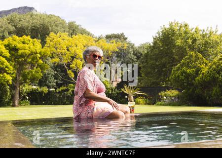 Femme biraciale senior profite d'un moment au bord de la piscine, ses cheveux gris complétant la journée ensoleillée Banque D'Images