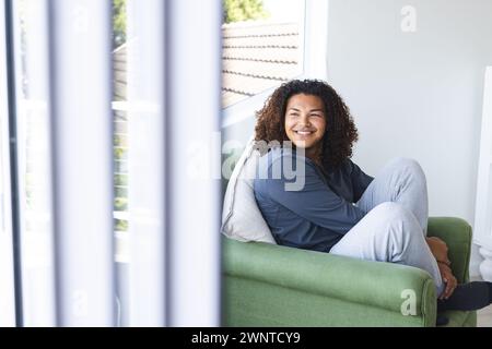 Jeune homme biracial avec les cheveux bouclés sourit tout en étant assis sur une chaise verte avec espace de copie Banque D'Images