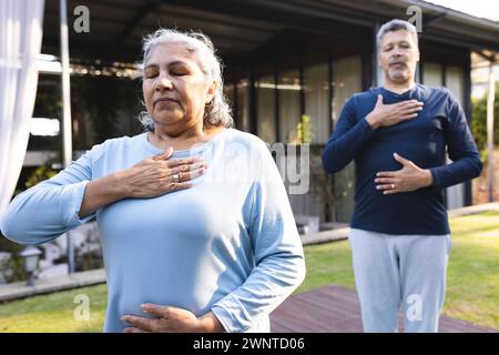 Femme biraciale senior et homme pratiquent le yoga à la maison, debout avec les yeux fermés et les mains sur les poitrines Banque D'Images