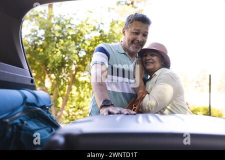 Couple senior biracial embrassant à côté d'un coffre de voiture ouvert, prêt pour un voyage sur la route Banque D'Images