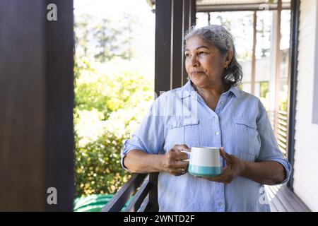 Femme biraciale senior avec les cheveux gris se tient sur un porche, tenant une tasse, avec un espace de copie Banque D'Images