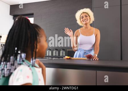 Une jeune mère biraciale fait des vagues à une fille afro-américaine dans une cuisine, allant à l'école Banque D'Images