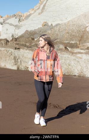 femme portant une veste patchwork et un pantalon noir marche sur une plage. La veste est colorée et a un design patchwork Banque D'Images