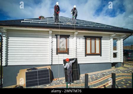 Ouvriers soulevant le panneau solaire sur un toit de maison. Trois ouvriers masculins portant des casques et des combinaisons de travail portant une cellule solaire. Concept de ressources énergétiques alternatives. Banque D'Images