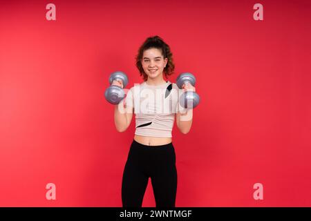 Adolescente sportive exerce avec des haltères pour développer des muscles isolés sur fond rouge. Banque D'Images