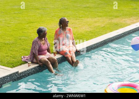 Deux femmes profitent d’une journée ensoleillée au bord de la piscine, les jambes trempées dans l’eau. Banque D'Images