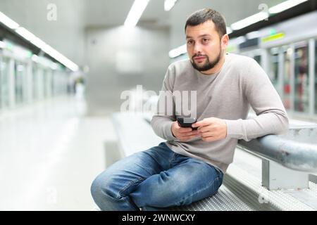 Homme barbu assis avec le téléphone sur le banc sur la plate-forme de métro Banque D'Images