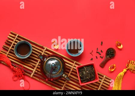Tapis de bambou avec théière, tasses de thé, bol et cuillère avec des feuilles sèches sur fond rouge Banque D'Images