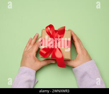 Main de femme tenant une boîte cadeau enveloppée dans un ruban de soie rouge sur un fond vert, vue de dessus Banque D'Images