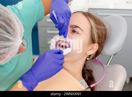 Conception des soins dentaires. Le processus d'installation des équerres orthodontiques en céramique. Vue rapprochée des mains du dentiste appliquant de la colle bleue sur la propreté du patient Banque D'Images