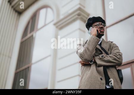 Un jeune homme d'affaires bien habillé avec des lunettes et un béret parle sur son téléphone intelligent avec une expression préoccupée, debout contre un bâtiment. Banque D'Images