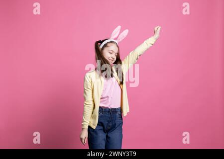 Jeune fille enthousiaste positive trompant autour et dansant sur la caméra, portant des oreilles de lapin en studio et montrant des mouvements de danse. Jeune enfant agissant joyeux et excité par la célébration de pâques. Banque D'Images