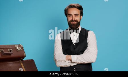 Concierge professionnel de l'hôtel posant à la caméra sur fond bleu, se préparant à transporter des bagages et à aider les voyageurs. Employé qualifié élégant travaillant comme chasseur, concept de l'industrie du tourisme. Banque D'Images