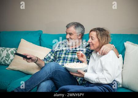 Dans le confort de leur salon, un couple sénior se détend sur le canapé, immergé dans une routine tranquille du soir. Avec la télécommande du téléviseur dans une main et un Banque D'Images