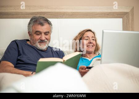 Dans cette charmante scène, un couple de personnes âgées profite de leur soirée ensemble au lit. On est plongé dans le monde numérique, travaillant sur un ordinateur portable, tandis que le Banque D'Images