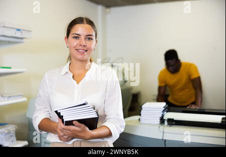 Portrait d'une employée amicale de la maison d'impression avec pile de carnets dans ses mains Banque D'Images