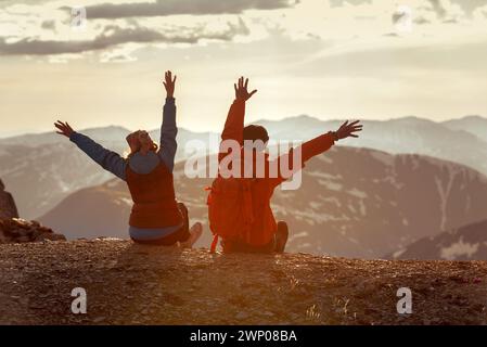 Deux randonneurs heureux avec des sacs à dos sont assis au sommet de la montagne à bras ouverts et profiter du coucher du soleil Banque D'Images