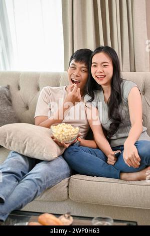 Joyeux et heureux couple asiatique rient, apprécient regarder un film et mangent du pop-corn sur un canapé à la maison ensemble. vie domestique, mari et wif Banque D'Images