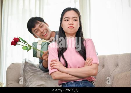 Un mari asiatique romantique essaie de demander pardon à sa femme en colère en la surprenant avec un cadeau et des fleurs dans le salon. famille Banque D'Images