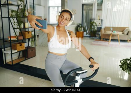 Femme hispanique dans des écouteurs prenant selfie lors du vélo d'exercice à la maison Banque D'Images