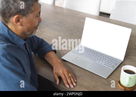 Homme senior biracial avec des cheveux grisonnants travaille sur un ordinateur portable à une table en bois, avec espace de copie Banque D'Images