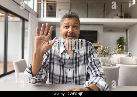 Homme senior biracial avec les cheveux gris vagues bonjour d'une cuisine moderne sur un appel vidéo Banque D'Images