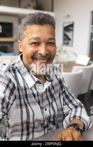 Homme senior biracial aux cheveux gris sourit chaleureusement lors d'un appel vidéo, portant une chemise à carreaux et une montre Banque D'Images
