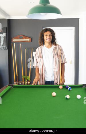Jeune homme biracial aux cheveux bouclés se tient près d'une table de billard, souriant Banque D'Images