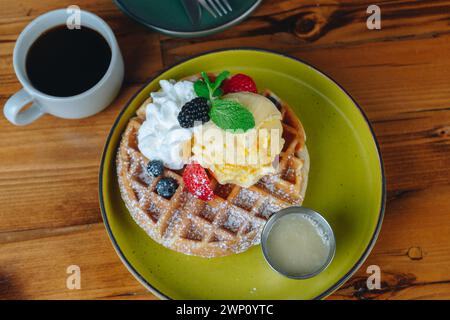 Une assiette de gaufres avec de la crème fouettée et des fruits sur le dessus se trouve sur une table à côté d'une tasse de café Banque D'Images