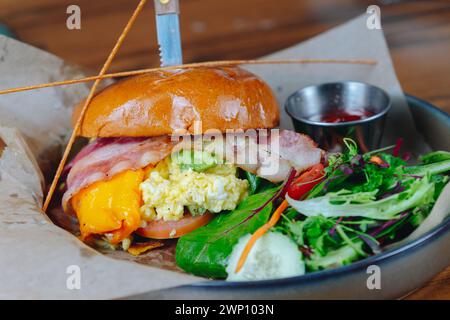 Un sandwich bacon et oeuf avec un accompagnement de salade. Le sandwich est sur une assiette avec un couteau qui en sort Banque D'Images