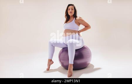 Jeune femme pratique le yoga de grossesse sur un ballon de stabilité dans un studio. Vêtue de vêtements de fitness, elle met en valeur sa bosse de bébé avec force et flexion Banque D'Images