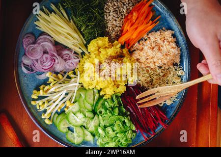 Une personne mange une salade avec une variété de légumes, y compris des carottes, du brocoli et des oignons. La salade est servie sur une assiette bleue et est placée dessus Banque D'Images