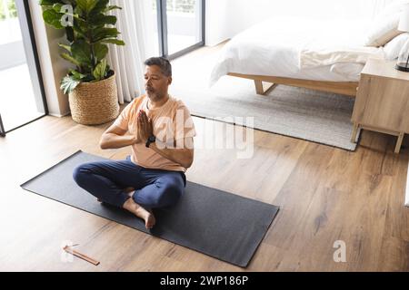 Homme senior biracial médite sur un tapis de yoga dans une chambre lumineuse, avec espace copie Banque D'Images