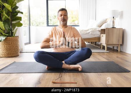 Homme senior biracial médite avec les mains en position de prière, assis sur un tapis de yoga Banque D'Images