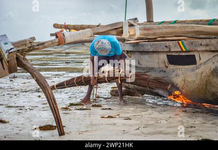 Un pêcheur utilise des frondes de palmier brûlantes pour ébavurer et imperméabiliser la coque de son bateau traditionnel à boutre en bois, Jambiani, Zanzibar, Tanzanie Banque D'Images