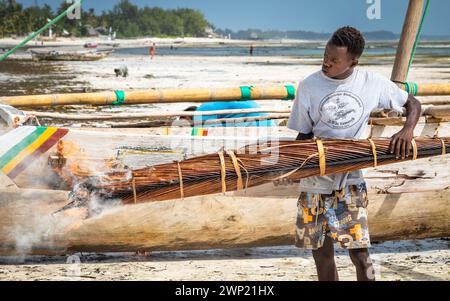 Un homme utilise des frondes de palmier brûlantes pour ébavurer et imperméabiliser son bateau traditionnel à boutre en bois, Jambiani, Zanzibar, Tanzanie Banque D'Images