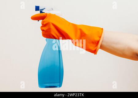 Une personne portant un gant en caoutchouc orange tenant un vaporisateur bleu transparent sur un fond Uni illustrant une action de nettoyage ou un concept d'hygiène Banque D'Images