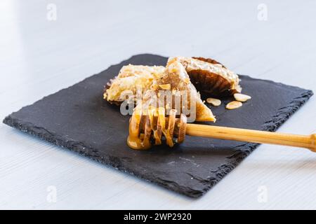 Photo horizontale une sélection de bonbons arabes glacés au miel ornés de noix, élégamment servis sur un plateau en pierre sombre, avec une trempette de miel en bois Banque D'Images