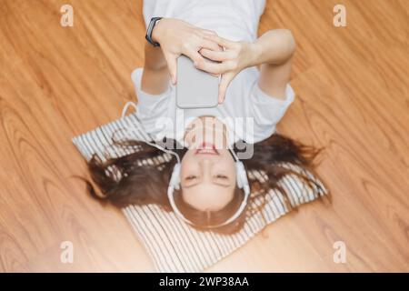 Vue de dessus d'une jeune femme asiatique magnifique avec casque et musique depuis un smartphone tout en étant couché sur le sol à la maison. Banque D'Images