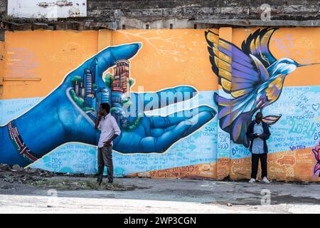 Deux hommes se tiennent devant des peintures murales représentant une main tenant une ville et un oiseau volant peint sur un mur dans la ville de Nakuru, créé dans le cadre du projet de ville créative de l'UNESCO de Nakuru pour mettre en valeur le patrimoine culturel et artistique de la ville. Ce travail collaboratif met en vedette les contributions de divers artistes de graffiti dont Bankslave, Joel Bergner, Chelwek, Dante et Natasha Floortje. Banque D'Images