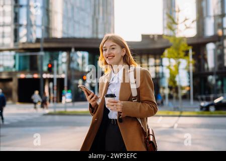Femme d'affaires confiante marchant avec son téléphone et son café le long du centre-ville Banque D'Images