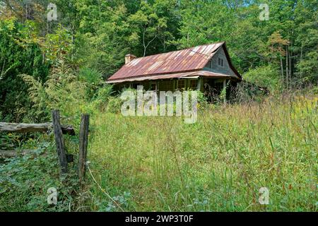 Une vieille maison vide en décomposition avec un toit en métal rouillé sur une colline dans les montagnes entourée d'arbres de mauvaises herbes envahis par la végétation et une vieille clôture en bois sur un soleil Banque D'Images