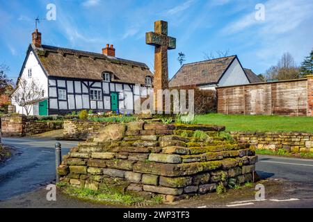 Croix en pierre dans le pittoresque village pittoresque du Cheshire d'Eaton by Tarporley avec un chalet noir et blanc à colombages de chaume Banque D'Images