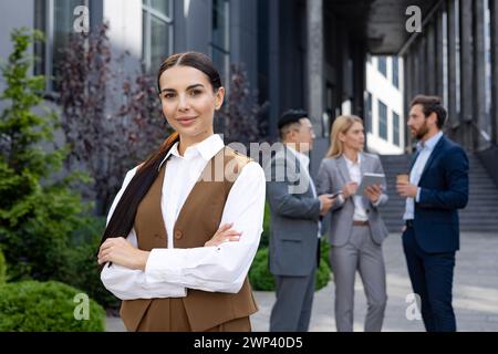 Femme professionnelle dans une tenue décontractée élégante posant en toute confiance avec les bras croisés. Collègues conversant en toile de fond. Banque D'Images