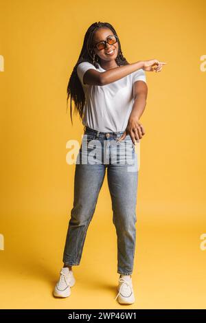 Vue pleine longueur de la femme enthousiaste excitée dans des lunettes de soleil dansant isolé sur jaune. Folle femme ravie en pull rose levant les mains, se déplaçant en rythmes. Bonheur et concept de style de vie Banque D'Images