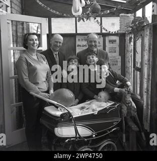 Années 1960, historique, grands sourires tout autour comme une famille debout pour une photo à l'intérieur d'un arrière-maison penché avec un enfant en bas âge assis dans une voiture d'entraînement construit tenant un ballon, Angleterre, Royaume-Uni. Banderole d'anniversaire au plafond. Banque D'Images